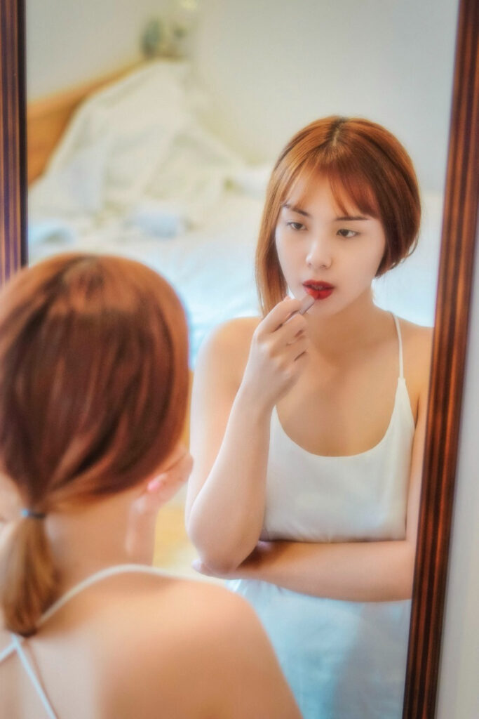 Girl looking at herself in the mirror, while putting red lipstick on her lips. She is showing concern on her face like she is not beautiful enough.