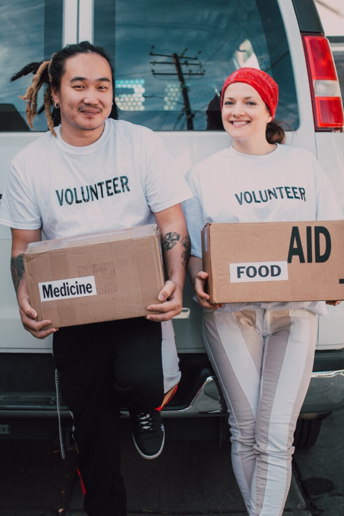 Woman and Man volunteering at a food drive holding boxes full of food and smiling, in front of a white van. The woman is showing true inner beauty.
