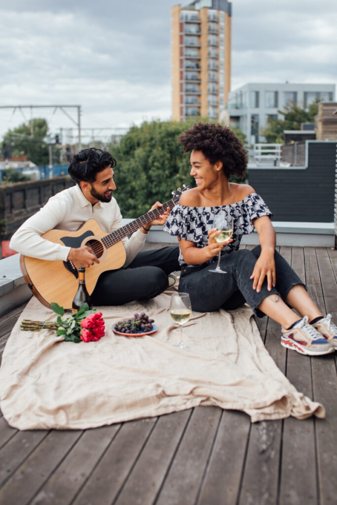 A captivating scene captured in a photograph, featuring a couple enjoying a romantic picnic on a rooftop in the city. The couple is seated on a blanket, surrounded by delectable food and drinks. The man lovingly strums a guitar, serenading his girlfriend, embodying the love language of acts of service and quality time.